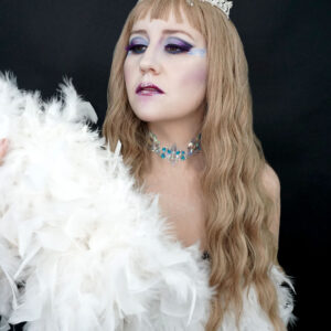 Mujer pálida rubia de cabello largo y ondulado vestida con un vestido blanco de plumas, maquillaje de colores fríos un, con una gargantilla y una corona