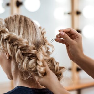 Se ve una chica con el color de pelo rubio, a la que le están habiendo un trenzado con moño para un boda