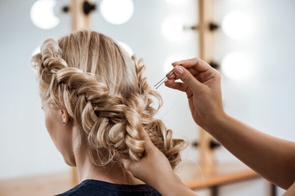 Se ve una chica con el color de pelo rubio, a la que le están habiendo un trenzado con moño para un boda